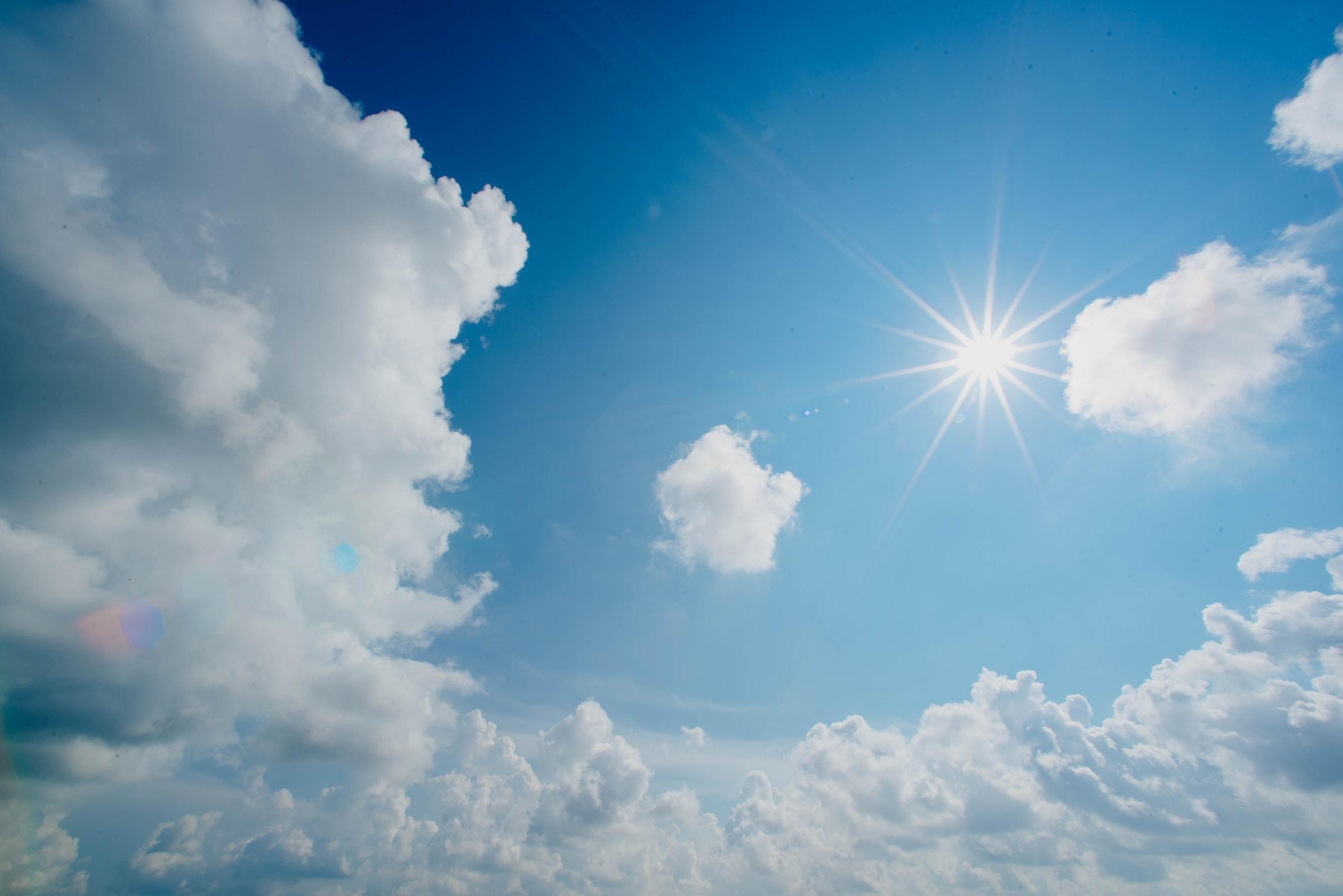 In einem blau-weiß bewölktem Himmel scheint die Sonne und wirft Strahlen wie ein Stern.