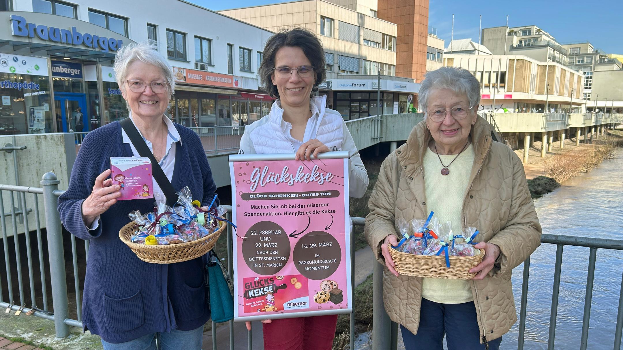 Drei Frauen stehen auf einer Brücke in der Neunkircher Innenstadt. Zwei von ihnen halten je einen Korb mit bunt verpackten Glückskeksen. Die Frau in der Mitte hält ein großes, pinkfarbenes Plakat mit Informationen zu einer Glückskeks‑Aktion. Im Hintergrund sind Geschäfte, Gebäude und die Blies zu sehen.