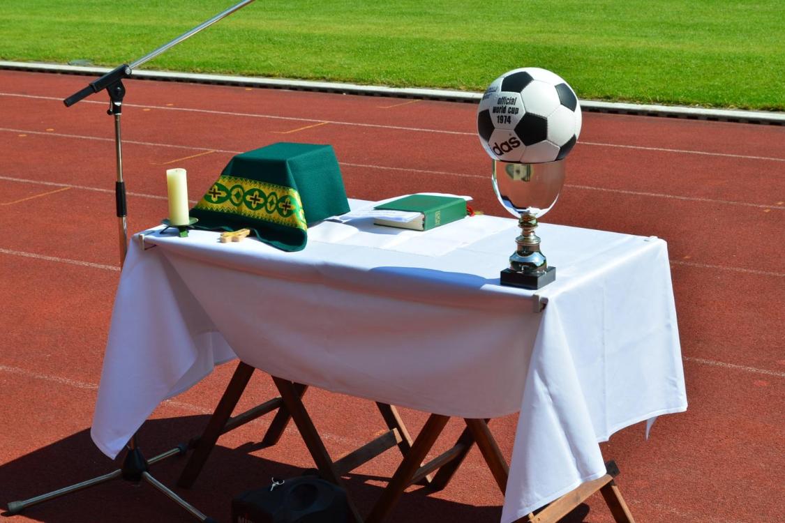 Vor dem grünen Rasen eines Stadions ist auf der roten Laufbahn ein provisorischer Altar aufgebaut. Auf dem weiß gedeckten Tisch liegen ein grünes liturgisches Tuch, eine Kerze, ein kleines Holzkreuz und ein liturgisches Buch. Links steht ein Mikrofonständer für eine Freiluftandacht oder einen Gottesdienst. Rechts auf dem Tisch befindet sich ein silberner Pokal, auf dem ein Fußball mit der Aufschrift „Official World Cup 1974“ platziert ist. Die Szene verbindet christliche Symbole mit dem Umfeld eines Sportplatzes.