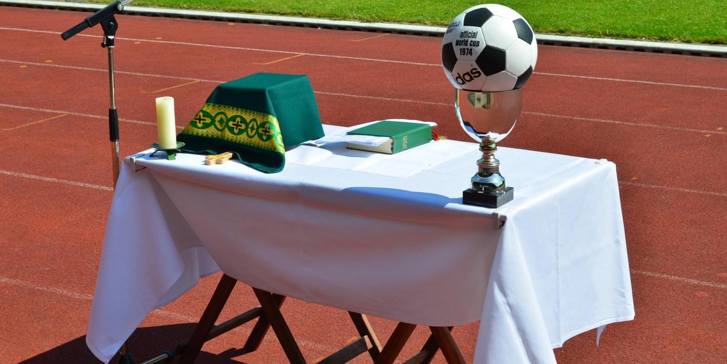 Vor dem grünen Rasen eines Stadions ist auf der roten Laufbahn ein provisorischer Altar aufgebaut. Auf dem weiß gedeckten Tisch liegen ein grünes liturgisches Tuch, eine Kerze, ein kleines Holzkreuz und ein liturgisches Buch. Links steht ein Mikrofonständer für eine Freiluftandacht oder einen Gottesdienst. Rechts auf dem Tisch befindet sich ein silberner Pokal, auf dem ein Fußball mit der Aufschrift „Official World Cup 1974“ platziert ist. Die Szene verbindet christliche Symbole mit dem Umfeld eines Sportplatzes.